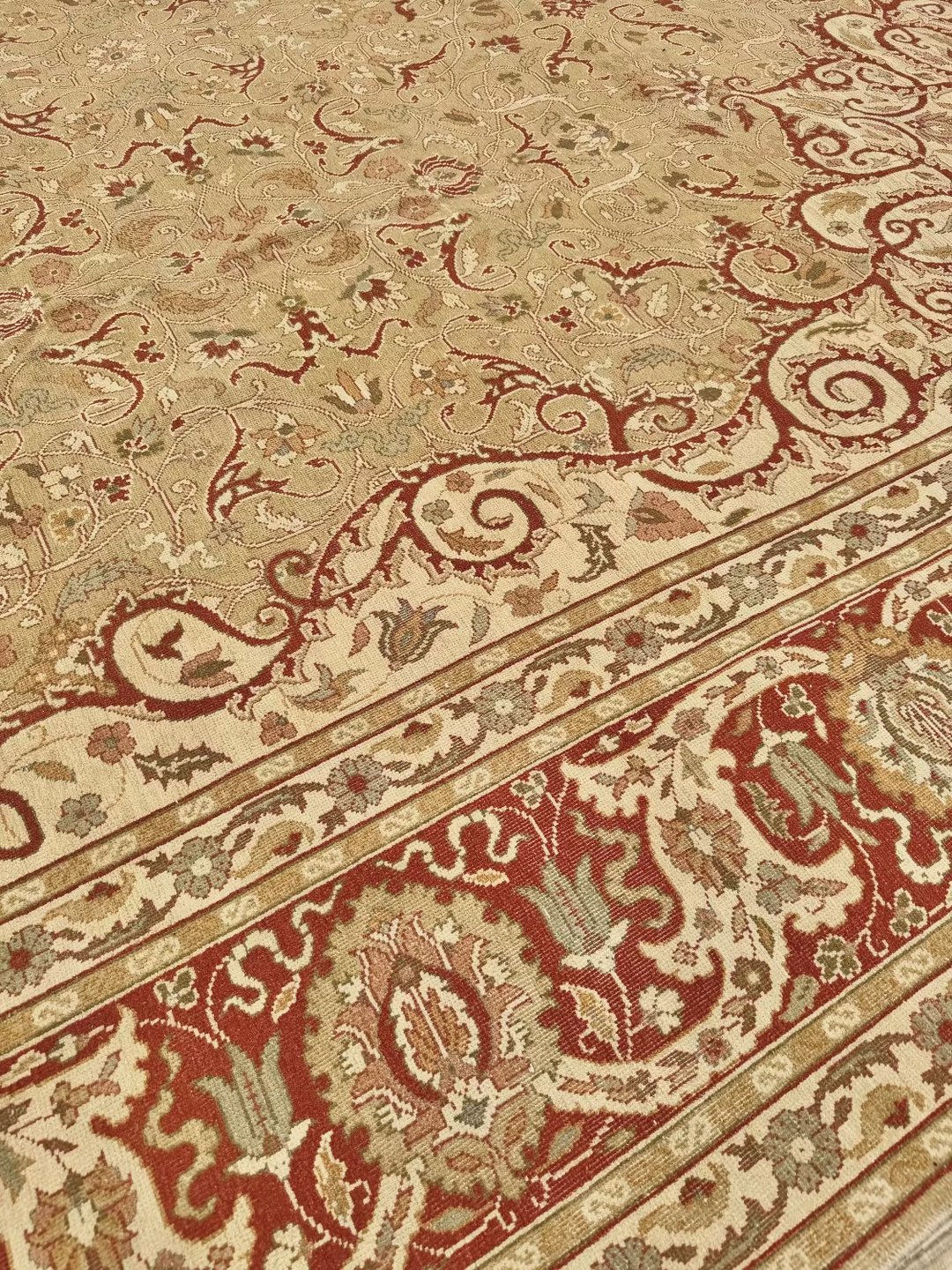 A close-up of an ornate area rug with a beige field, deep red borders, and intricate floral and scroll motifs in cream, rust, and muted green.