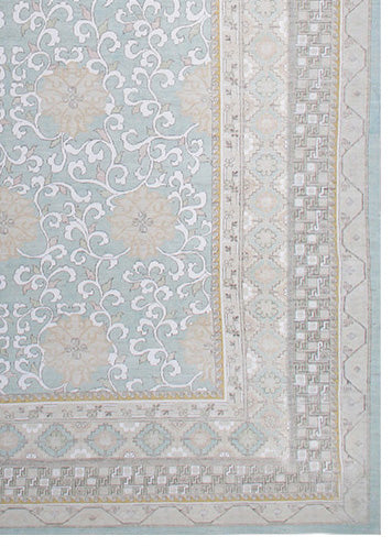 A close-up of an ornate patterned rug showing a light blue field with white scrolling floral motifs and beige borders.